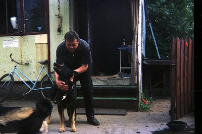 A man in a black shirt kneels interacting with a German Shepherd dog in a courtyard setting. A vintage blue bicycle and weath...