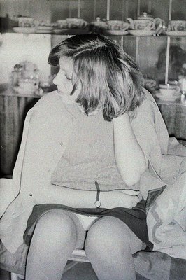 A young woman sits pensively in what appears to be a kitchen, gazing downwards. She wears a belted tunic and dark skirt. Back...