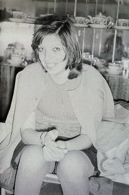 A young woman with short, dark hair sits casually, appearing relaxed. She wears a short-sleeved top and dark shorts. Behind h...
