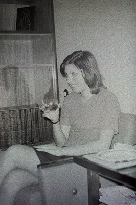 A young woman sits at a table, holding a stemmed glass, appearing to be in conversation. Likely a candid moment, the interior...