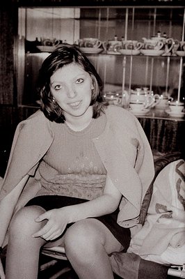 A young woman sits facing the camera, a slight smile on her face. She wears a short-sleeved patterned top and what appears to...