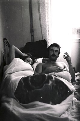 A couple rests in a bed, the man smoking and appearing relaxed, while the woman has her eyes closed. The photograph's grainy ...