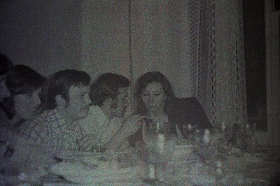 Group seated around a table for a gathering. Visible are five individuals with 1970s hairstyles, enjoying a meal. A lace curt...