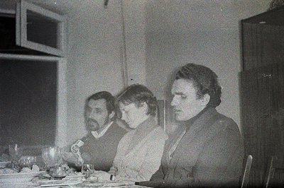 Three individuals seated at a table set for a meal. Likely a candid, domestic scene. Men display dark, full beards and short ...