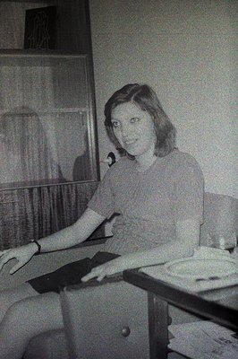 A young woman sits at a table, looking towards the camera. She wears a short-sleeved top and has a bracelet on her left wrist...