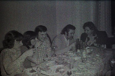 Five individuals gathered around a table laden with food and drink, possibly during a meal. The subjects display 1970s hairst...