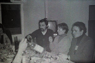 A black and white photo captures a group gathered around a laden table, possibly during a meal. Three men are seated prominen...