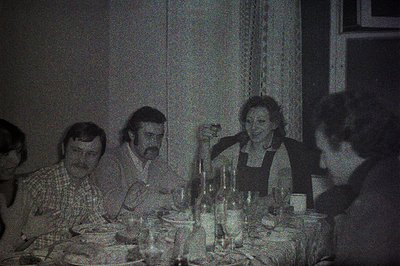 A candid, grainy, black-and-white photograph captures a family gathering at a dining table. Four individuals are seated, enga...