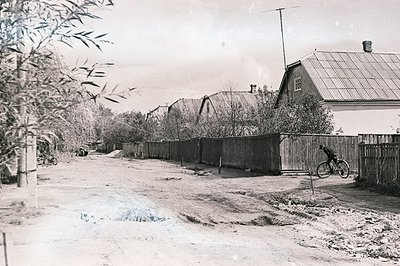 A dirt road winds between modest, gabled houses with corrugated metal roofs. A man cycles past a tall, weathered wooden fence...