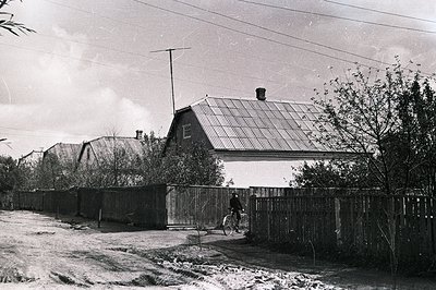 A low-angle view of a rural dwelling with a corrugated iron roof and brick chimney. A single cyclist rides along a narrow pat...