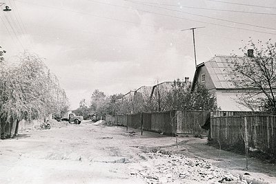 A snow-covered street scene depicts modest homes with steeply pitched roofs and wooden fences. A vintage truck is parked mid-...