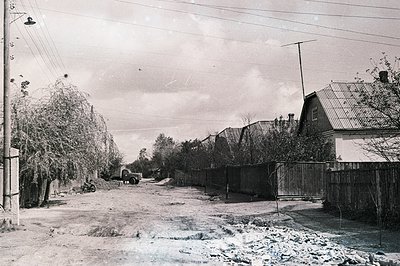 Dusty, unpaved road runs between aging homes and a weathered wooden fence. A vintage truck sits mid-street; sparse vegetation...