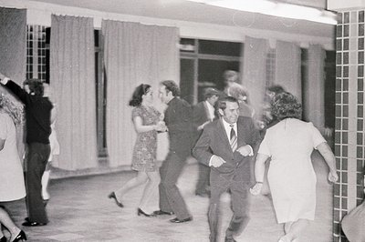 Energetic group dancing in a tiled interior, likely a hotel or event space. Individuals in 1960s attire - short dresses, suit...