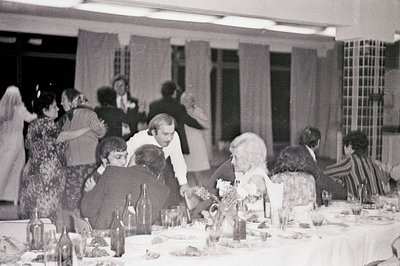 A lively banquet scene: guests seated at a long table with ornate floral centerpiece, celebrating. Men in suits, women in dre...