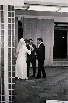 A formally attired couple, likely a bride and groom, stand close in what appears to be an indoor courtyard. The bride wears a...