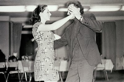 A couple dances in what appears to be a banquet hall or community center. The woman wears a short, patterned dress; the man, ...