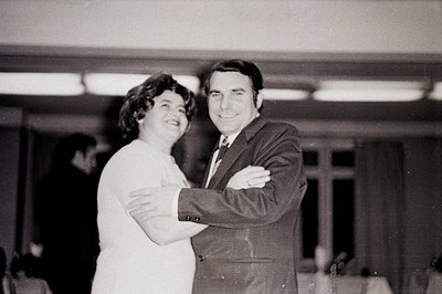 A couple in formal attire embrace in a warmly lit interior. The woman wears a long-sleeved dress, her hair dark and curly. Th...
