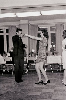A couple dances in a formal setting. Man in a dark suit, woman in a short, patterned dress and heels. Tables with white table...