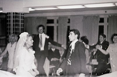 A candid wedding scene: a bride and groom dance, surrounded by formally dressed guests in a large, windowed room. Likely a 19...