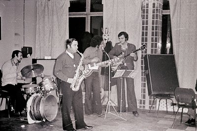 Black and white image depicts a jazz quartet rehearsing. A drummer sits behind a kit, while a saxophonist stands mid-performa...