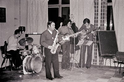 A black and white image captures a jazz quartet performing on a simple stage. A drummer sits on a stool, facing a drum kit, w...