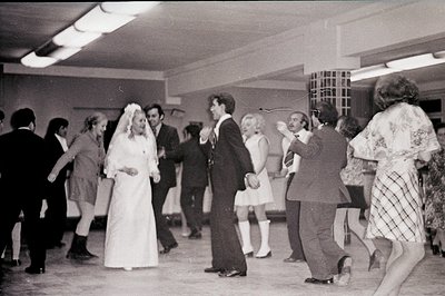 A candid wedding reception scene depicts a bride in a long-sleeved gown with a veil dancing with her groom in a dark suit. Gu...