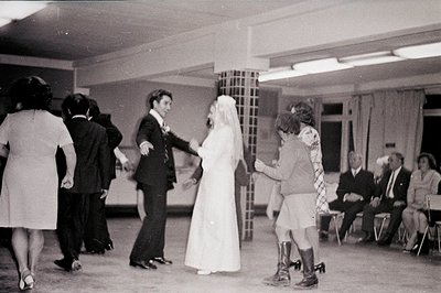 A bride and groom dance during a lively wedding reception, surrounded by guests in period attire (1970s). The scene takes pla...
