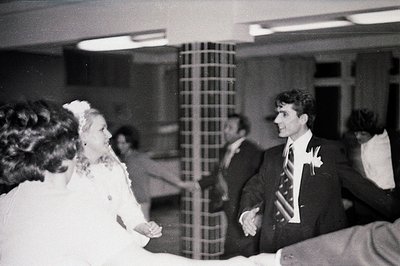 Black and white image depicting a wedding reception scene. A bride in a white gown and a groom in a dark suit stand facing ea...