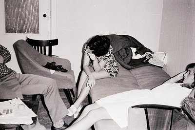 A candid snapshot depicts three individuals in a domestic interior, circa 1960s. Two figures recline on a sofa, while a third...