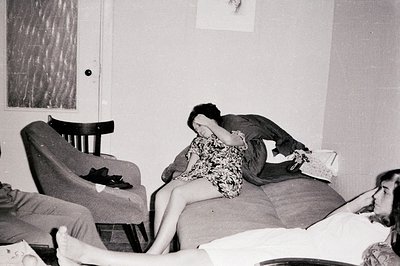 A woman in a patterned dress sits on a sofa, face buried in hands. Another woman lies on the floor, legs extended, displaying...