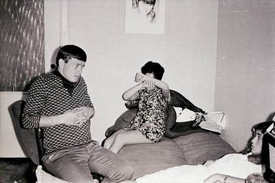 Three young adults, two male and one female, are seen in a sparsely furnished interior. The man sits, patterned sweater visib...
