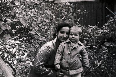 A woman holds a young child in her arms, both appearing warmly dressed. Behind them is a rustic wooden fence and lush foliage...