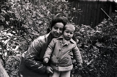 A young woman supports a small child in a warmly-dressed embrace, set against a backdrop of dense foliage and a weathered woo...