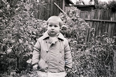 A young boy stands outdoors, framed by dense foliage and a weathered wooden fence. He wears a padded coat with double-breaste...