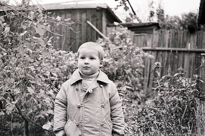 A young boy in a padded coat and turtleneck stands outdoors, framed by foliage and a weathered wooden fence. The image’s styl...