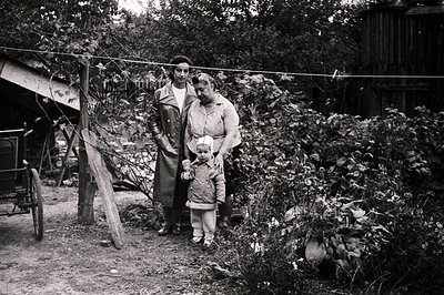 A family stands in a rural yard, likely post-war era (1950s-1960s). The mother, in a trench coat, holds the hand of a young b...