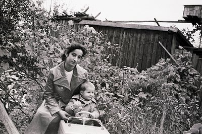 A young woman and toddler sit on a toy car in a backyard setting. A weathered, wooden fence and shed form the backdrop. The i...