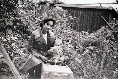 A young woman in a trench coat sits with a baby in a toy car, posed in front of a weathered wooden fence and lush foliage. Li...