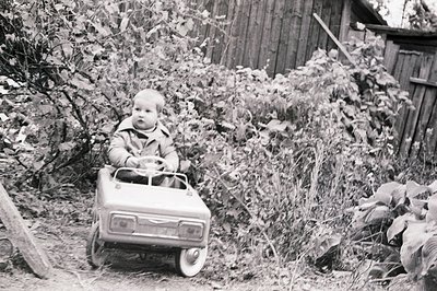 A young boy sits in a vintage pedal car toy, surrounded by lush foliage and a weathered wooden fence. The car's design evokes...