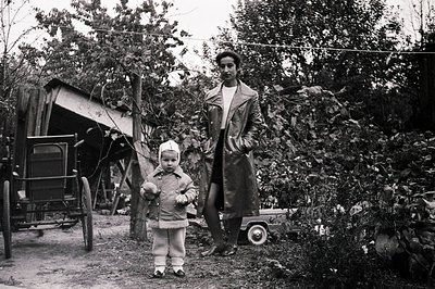 A young child bundled in a hooded coat stands before a tall figure in a long coat, near a dilapidated shed and overgrown foli...