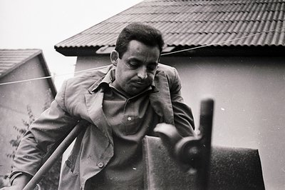 Man in a light-colored suit leans on a balcony railing, gazing downwards. Close-up framing emphasizes his face and the textur...