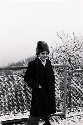 A young person stands outdoors in winter clothing, likely a fur hat, dark coat & trousers. A snow-covered landscape forms the...