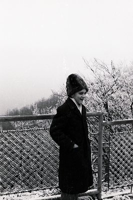A young woman stands outdoors in a heavy winter coat and fur hat, looking toward the horizon. The snowy landscape suggests a ...
