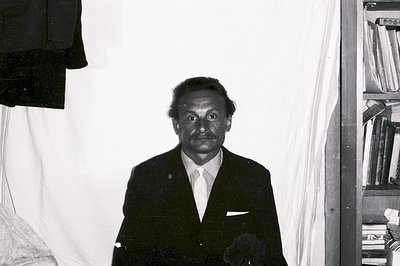 Formal portrait of a man in a suit and tie, posing within a room. Visible are a draped cloth, and a stacked bookshelf. Minima...