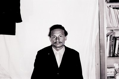 Striking portrait of a man in a dark suit and tie, posed formally against a plain backdrop. Noticeable dark circles around ey...