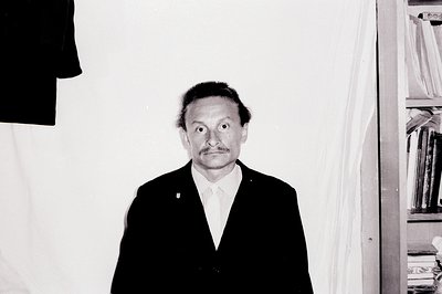 A formal portrait of a man in a dark suit and tie, facing forward with a subtly distressed expression. The background feature...