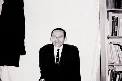 Man in a dark suit and tie stands near bookshelves, appearing startled or distressed. Black and white photo suggests a mid-ce...