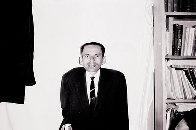 A black and white portrait of a formally dressed man standing near a bookcase. He wears a dark suit, tie, and appears slightl...