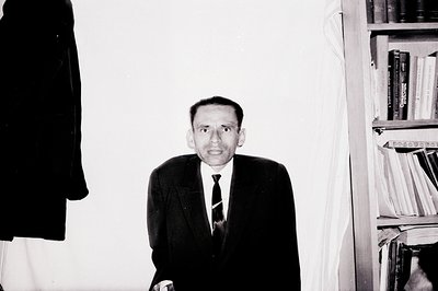 Man in a dark suit and tie stands before a bookshelf, appearing contemplative. The stark black and white contrasts emphasize ...