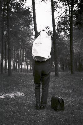 A solitary figure stands in a wooded area, obscured by a paper bag covering their head. Dark trousers and leather shoes are v...